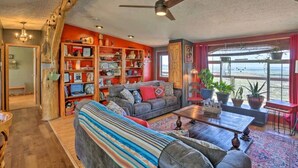 A cozy living room with a sofa, coffee table, and bookshelf. There is a large window with a view of the ocean. - Cozy Black Hills Home on 13 Acres w/ Deck & Views!! (Hot Springs)