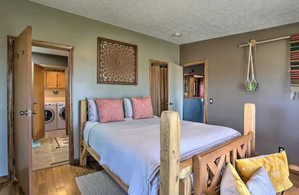 Une chambre à coucher comprenant un lit, une tête de lit en bois, une œuvre d’art accrochée au mur et une porte donnant sur une buanderie.