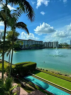 A large body of water with a fountain, surrounded by palm trees and a well-maintained lawn. In the background, there are high-rise buildings. - Marriott villa 2-bedroom in Doral Miami with all amenities! PGA GOLF EVENT week (Miami)