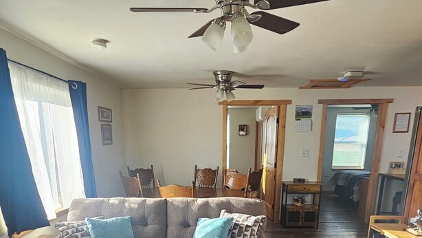 A living room with a sofa, ottoman, ceiling fan, wooden flooring, and a dining area with chairs and a table. - Mountain Crest Cabin - Cozy Lodging, Close to Cody (Cody)