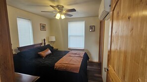 A bedroom with a bed, a ceiling fan, two windows with blinds, a nightstand with a lamp, and a wooden door. - Mountain Crest Cabin - Cozy Lodging, Close to Cody (Cody)