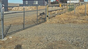 A fenced property with a gravel driveway, a metal gate, and a building with a dark roof. - Mountain Crest Cabin - Cozy Lodging, Close to Cody (Cody)