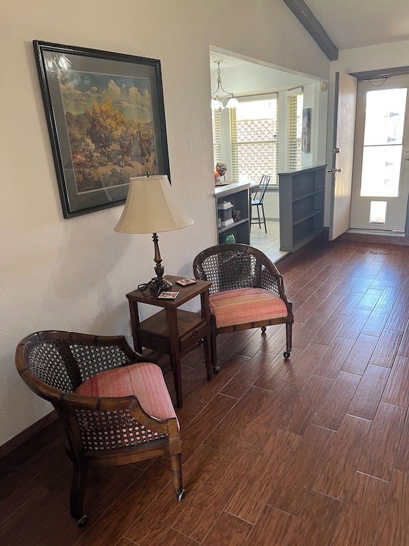A room with wooden flooring, two wicker chairs, a small table with a lamp, and a framed painting on the wall.