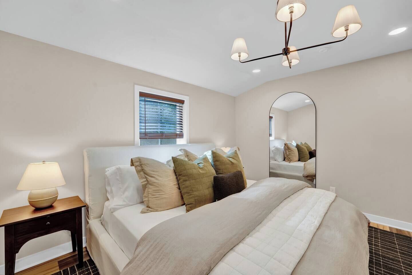 A hotel room with a bed, a bedside lamp, a window with blinds, a mirror, and a ceiling light fixture.