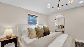 A hotel room with a bed, a bedside lamp, a window with blinds, a mirror, and a ceiling light fixture.