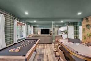 Uma mesa de bilhar com bolas, um sofá, uma televisão e um espaço de bar com petiscos.