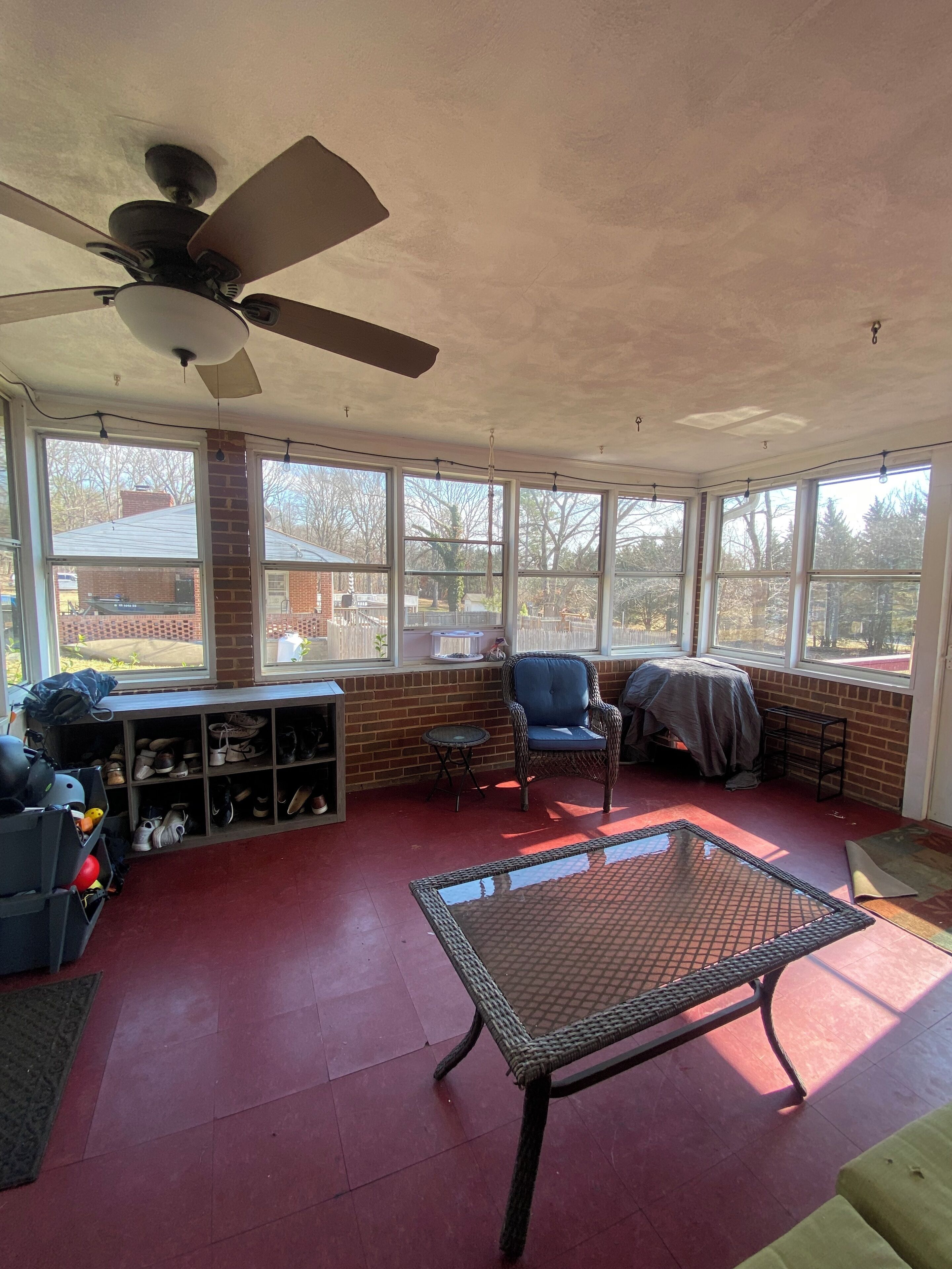 A sunroom with a ceiling fan, large windows, a wicker chair, a glass table, and a shoe rack.