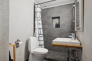 A bathroom with a toilet, shower curtain, tiled walls, a sink with a mirror, and a small shelf with toiletries.