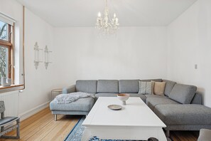 Un salon moderne avec un canapé d’angle gris, une table basse blanche, un lustre et une fenêtre équipée de stores.