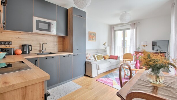 Een moderne keuken met grijze kastjes, een magnetron en een koffiezetapparaat. Er is een zithoek met een tafel, stoelen en een bloemenvaas.