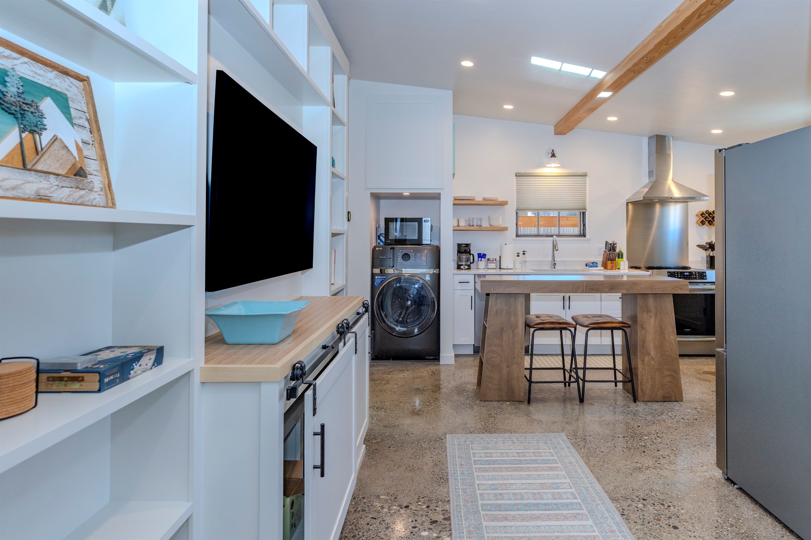 A modern kitchen with a built-in washing machine, a wooden dining table, and a TV mounted on the wall.
