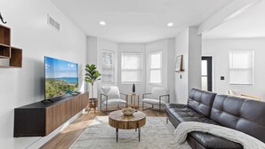 Ein modernes Wohnzimmer mit Flachbildfernseher, Couchtisch aus Holz, Ledersofa und zwei weißen Sesseln.