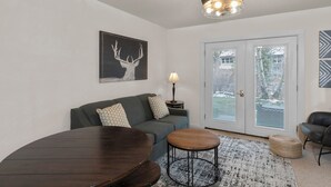 A modern living room with a dark wood dining table, a grey sofa with patterned cushions, a small round table, and a lamp.
