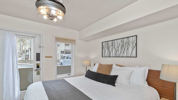 A hotel room with a large bed, a bedside table with a lamp, a window with a view of the street, and a door leading outside.