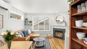 A modern living room with a fireplace, a dining area with a table and chairs, a sofa with cushions, and a bookshelf.