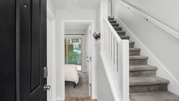 A hallway with a dark door, a staircase with grey carpeted steps, and a bed in the background.