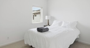 Ein sauberes, weißes Schlafzimmer mit einem ordentlich bezogenen Bett, einer Nachttischlampe und einem Fenster mit Jalousien.