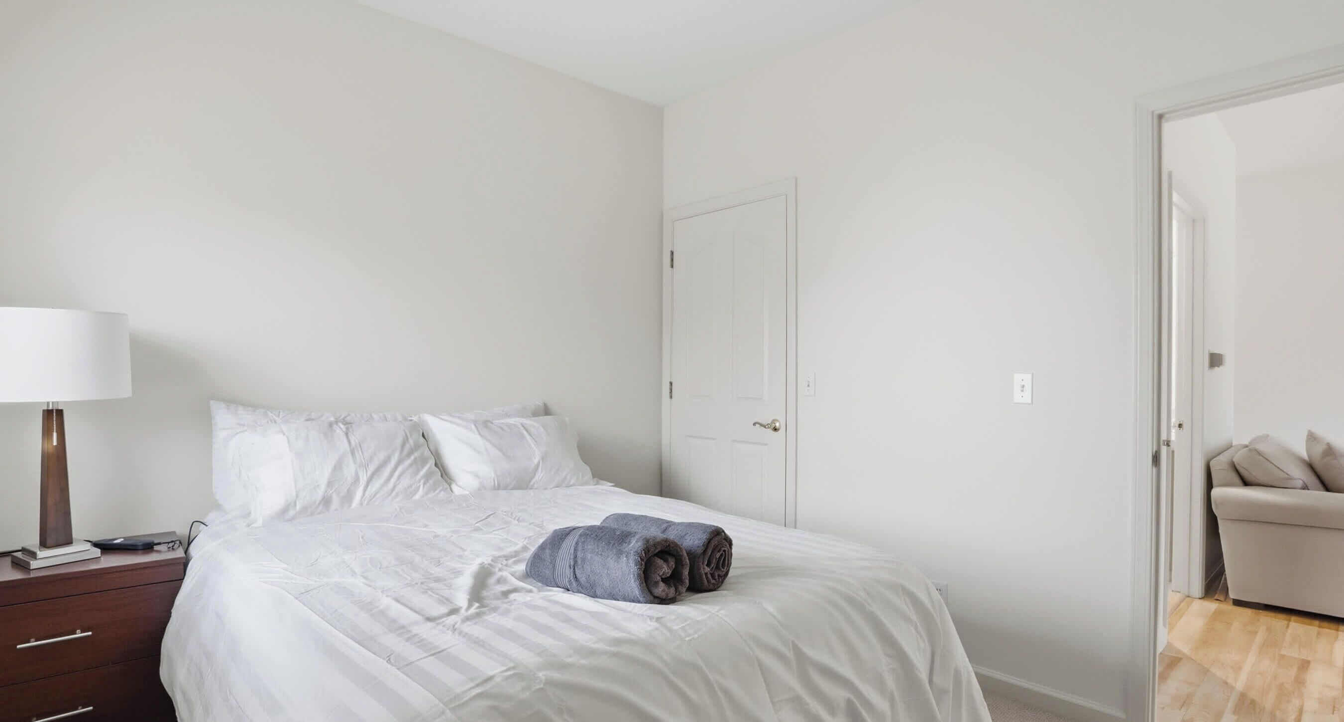 Een slaapkamer met een bed, een nachtkastje met een lamp, en een deur naar een andere kamer.