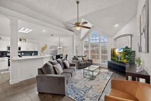 A spacious living area with a high ceiling, a ceiling fan, a large TV, and a kitchen in the background. - Upgraded Pool Home Minutes to UNT & Denton (Lake Dallas)