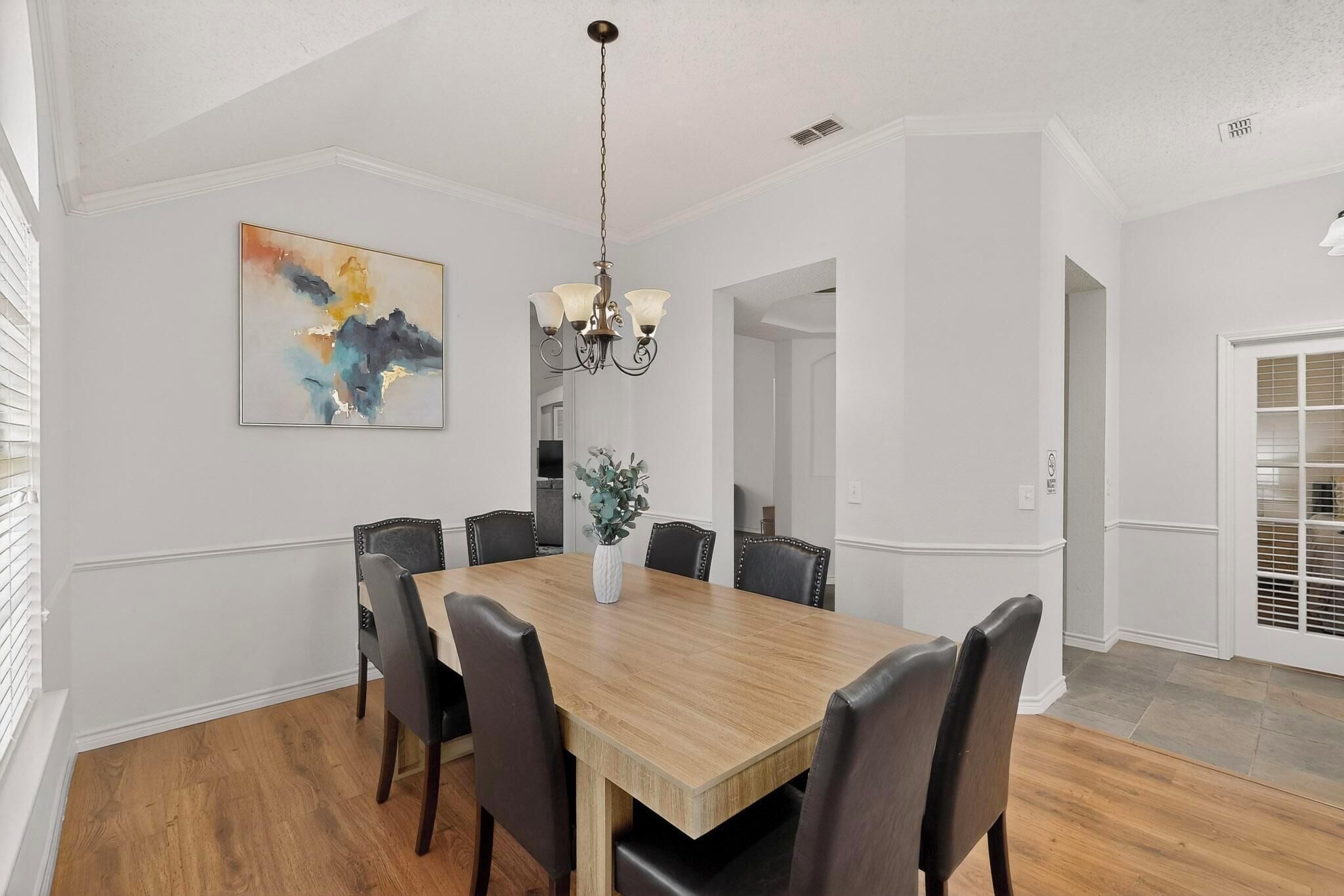 Un comedor con una mesa de madera, seis sillas, una araña y un cuadro en la pared.