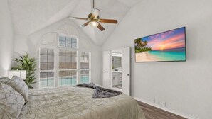 Une chambre avec un lit, un ventilateur de plafond, une grande fenêtre avec des stores, une porte et un téléviseur fixé au mur qui diffuse une scène de plage.
