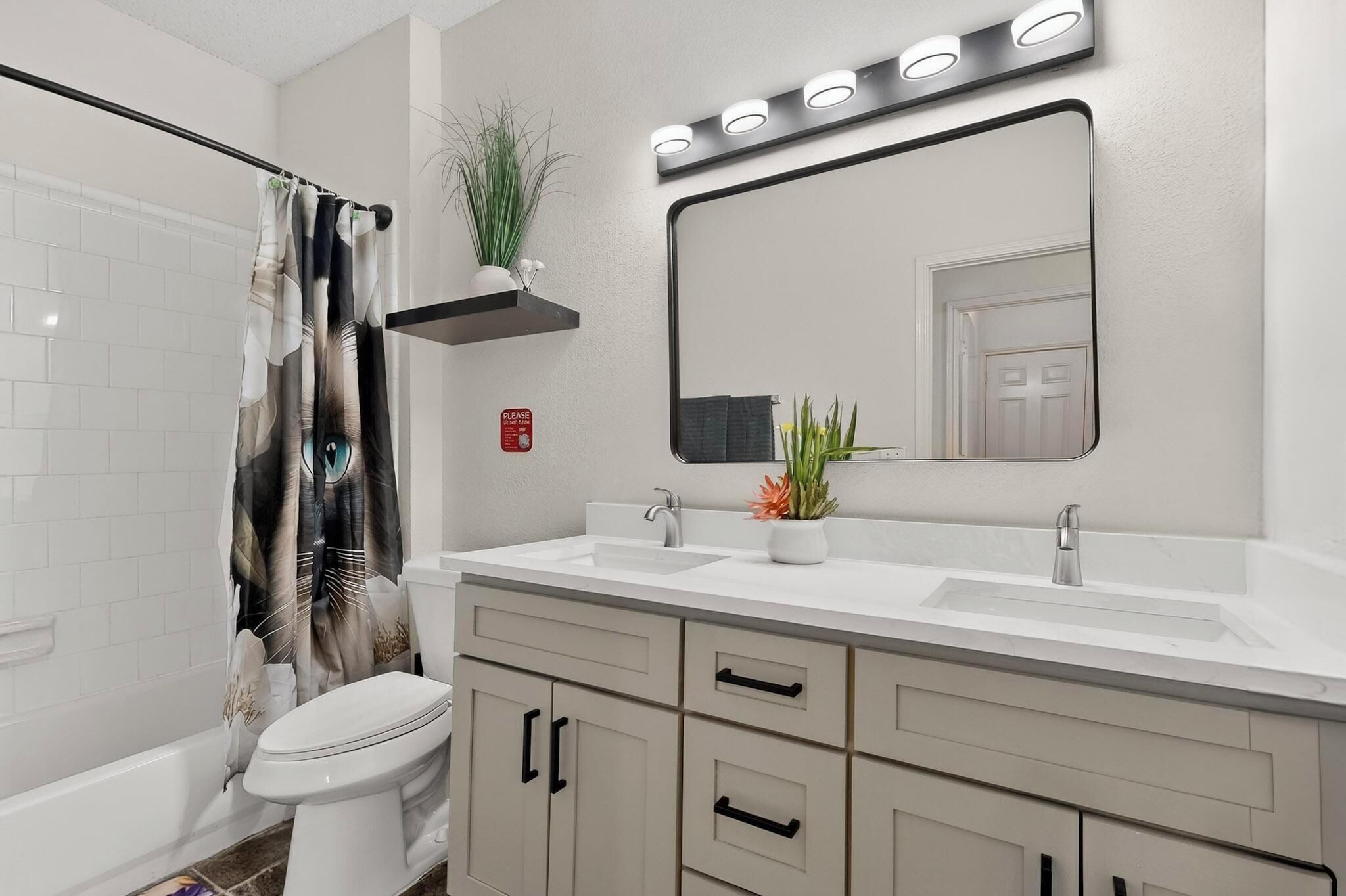 Ein Badezimmer mit Waschbecken, Spiegel und Toilette. Eine Duschvorhang mit Blättermotiv, eine kleine Pflanze auf einem Regal und ein Schild mit dem Schriftzug "Bitte" an der Wand.