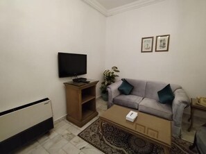 A living room with a flat-screen TV, a grey sofa with two cushions, a wooden side table, a coffee table, and a radiator. - Rotana Furnished Apartments (Jeddah)