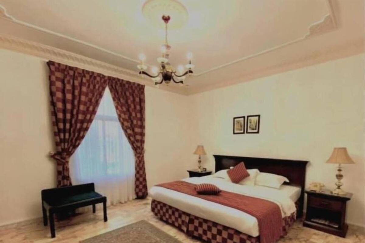 A bedroom with a large bed, a bench, a chandelier, and a rug.