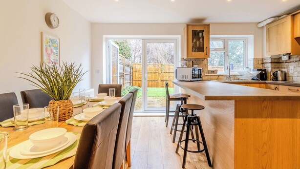Una cocina moderna con una isla central, comedor con una mesa preparada para cuatro personas y vista al patio trasero a través de puertas de vidrio.