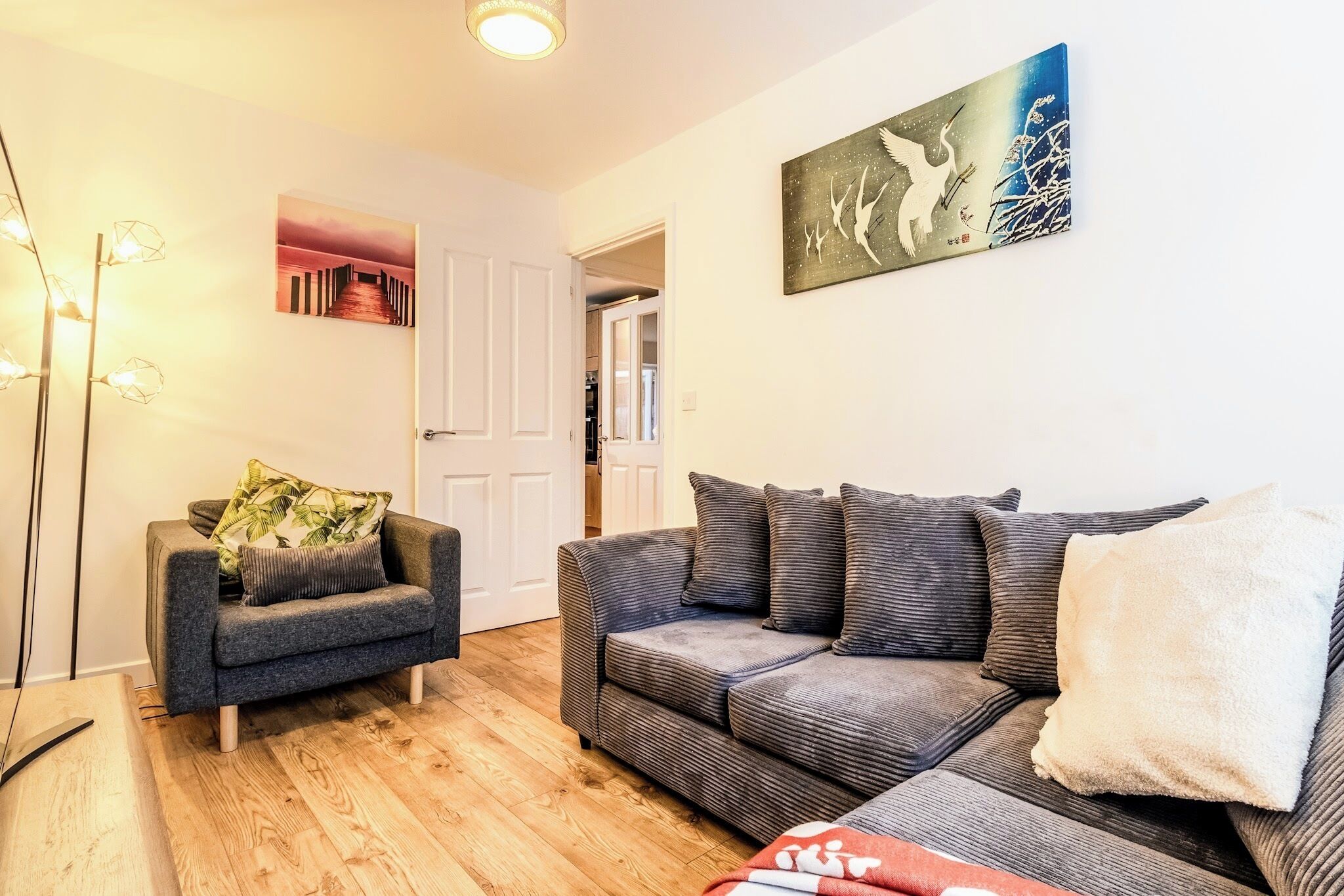 A living room with a grey sofa, a smaller armchair, and a wooden floor. There is a door leading to another room, and a painting on the wall.