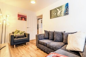 A living room with a grey sofa, a smaller armchair, and a wooden floor. There is a door leading to another room, and a painting on the wall. - Spacious 3 bedroom home, perfect for families & contractors. Free parking (Reading)