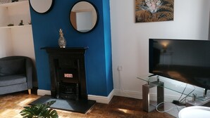 A living room with a blue wall, two round mirrors, a fireplace with a decorative item, a television on a glass stand, and a potted plant. - Ermine Road -  a house that sleeps 7 guests  in 4 bedrooms (Chester)