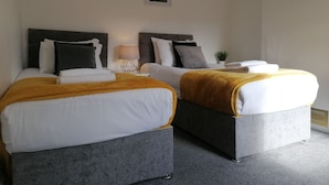 A double room with two beds, each with a grey headboard, white bedding, and a mustard-colored throw blanket. There is a bedside table with a lamp and a small potted plant. - Ermine Road -  a house that sleeps 7 guests  in 4 bedrooms (Chester)