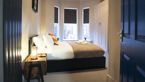 A bedroom with a bed, bedside table, lamp, and windows. - Brattan Road -  a house that sleeps 5 guests  in 5 bedrooms (Oxton)