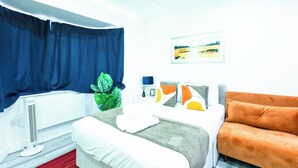 Ein Hotelzimmer mit Bett, einer Couch, einem Fenster mit Vorhängen, einem Heizkörper, einer Pflanze und einem Bild an der Wand.