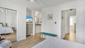 A bedroom with a bed, a bench, a ceiling fan, a TV, a kitchen area, and a bathroom. - Prime Location/2 Bdr/Sleeps 7/Pet friendly (Santa Rosa Beach)
