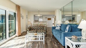 A spacious living room with a blue sofa, patterned armchair, and a white coffee table. A large window provides a view of the city. - Two oceanfront units, side by side. Perfect for extended family and golf group vacations (Myrtle Beach)