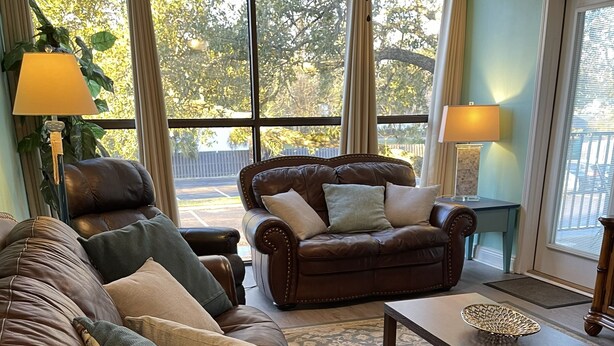Un salón con un sofá de cuero marrón, un sillón a juego y una pequeña mesa con un cuenco decorativo. Amplias ventanas ofrecen vistas a árboles y una valla.