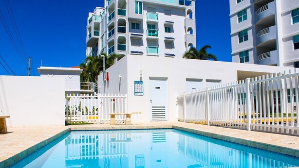 Une piscine entourée d’une clôture blanche, un banc au bord de la piscine et, au fond, un bâtiment doté de plusieurs balcons.