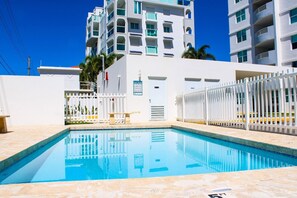 A swimming pool with a white fence, a poolside bench, and a building with multiple balconies in the background. - Beachfront Paradise in Rincón (Stella)