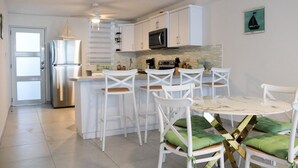 Une cuisine moderne avec des meubles blancs, un réfrigérateur en acier inoxydable, un four à micro-ondes et un four encastré. On y trouve des chaises blanches avec des coussins verts disposées autour d’une table en marbre.