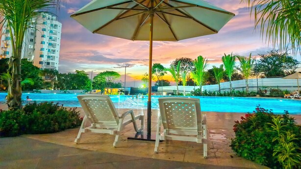 A poolside area with two white lounge chairs and a large umbrella, surrounded by palm trees and a building in the background. - Apartaloft Green Paradise - Santa Marina (Santa Marta)