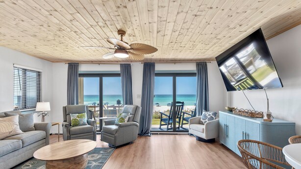 Ein großzügiges Wohnzimmer mit Holzbalkendecke, großen Fenstern, einem Flachbildfernseher und Blick auf den Strand.