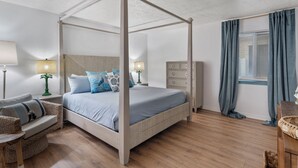 Ein Bett mit Baldachin in neutralen Farben, ein hölzerner Schminktisch, ein Nachttisch mit Lampe, ein Korbstuhl und ein Fenster mit Vorhängen.