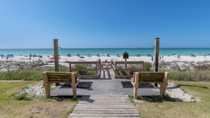 Ein hölzerner Weg führt zu Liegestühlen mit Blick auf einen Sandstrand, Sonnenschirme und Menschen.