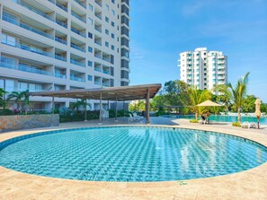 Ein Swimmingpool mit gefliestem Sonnendeck, umgeben von einer überdachten Sitzgelegenheit und Palmen.