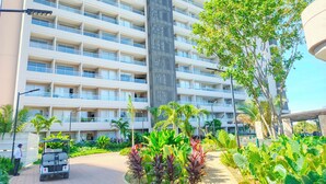 Un hôtel à plusieurs étages, doté d’une tour centrale, entouré d’une végétation luxuriante et accessible par une allée pavée.