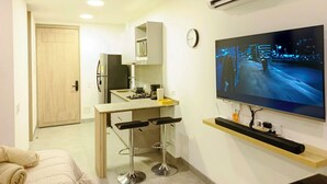 Un espace de vie compact comprenant un coin cuisine, un téléviseur fixé au mur et une porte donnant sur une autre pièce.