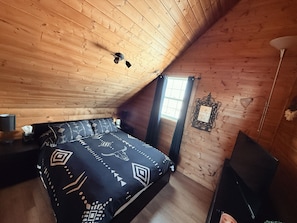 A bedroom with a sloped ceiling, a bed with patterned bedding, a bedside table with a lamp, a window with curtains, and a wall-mounted picture frame. - Mirrored Waters Ranch Cabin (Mirror)
