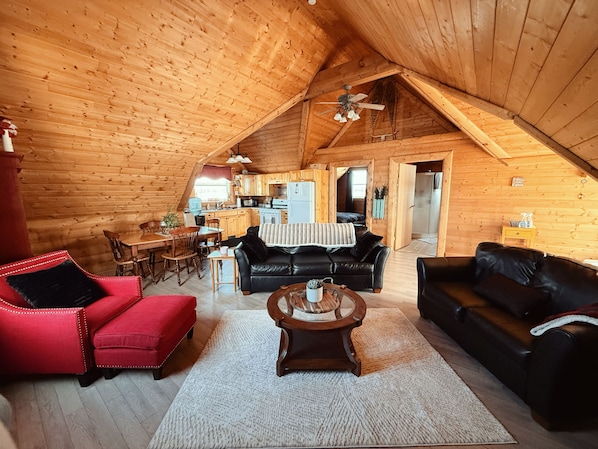 Interior de una cabaña de troncos con comedor, cocina y sala de estar, amueblada con sofás de cuero y un sillón rojo.